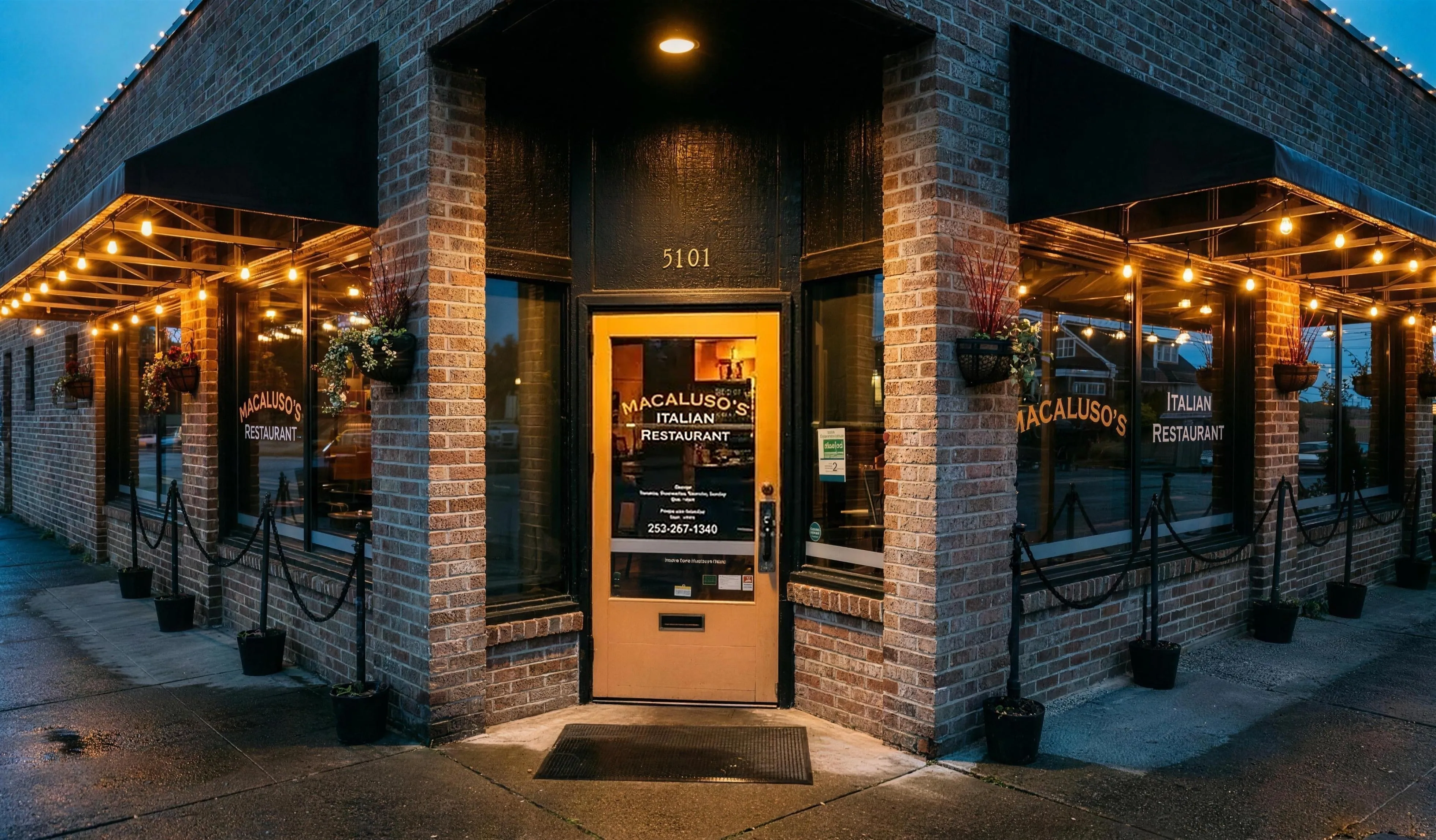 The Macaluso’s brick corner storefront at dusk, string lights glowing and the gold window lettering lit.