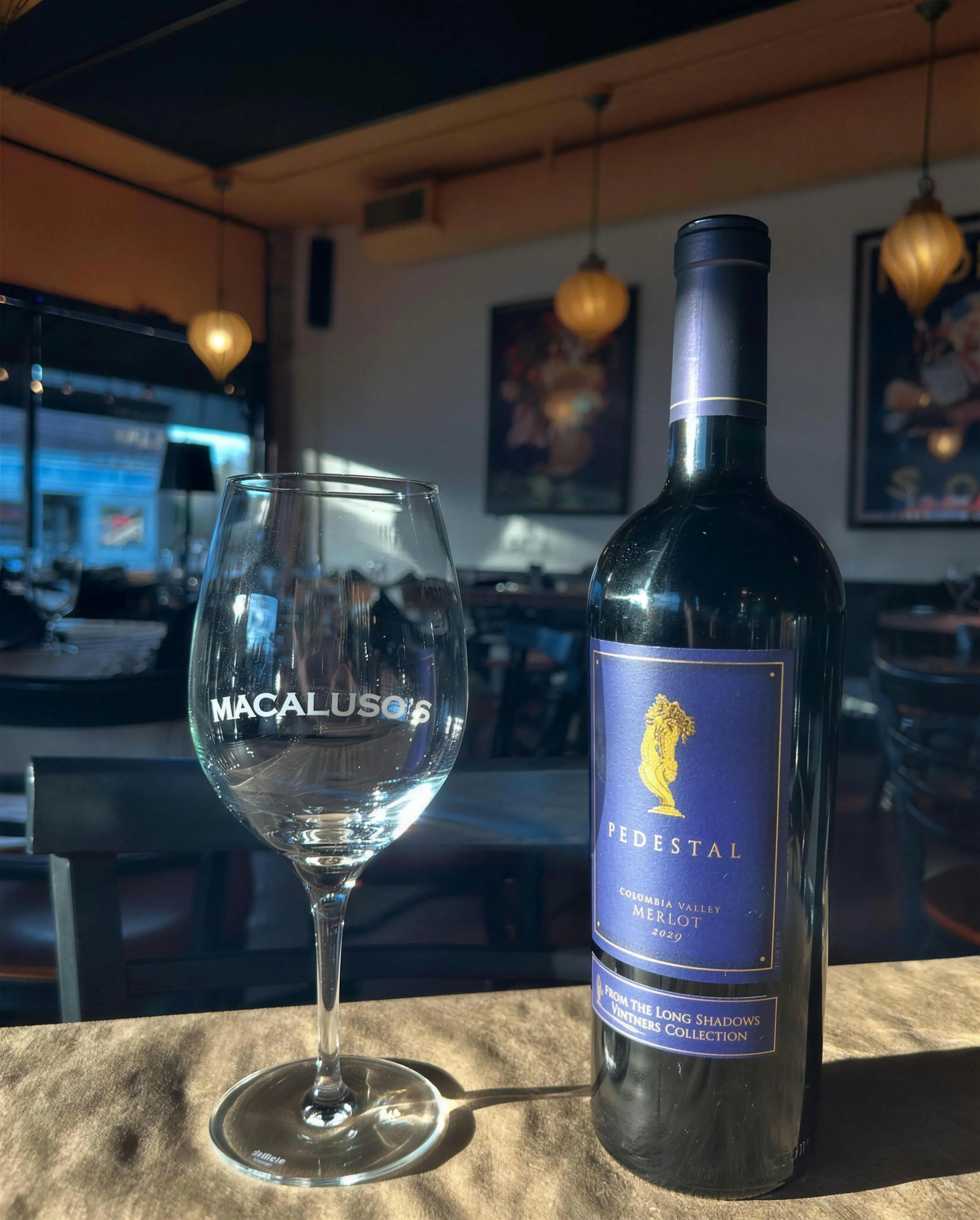 A Macaluso’s etched wine glass beside a bottle of Pedestal Merlot in a sunbeam.