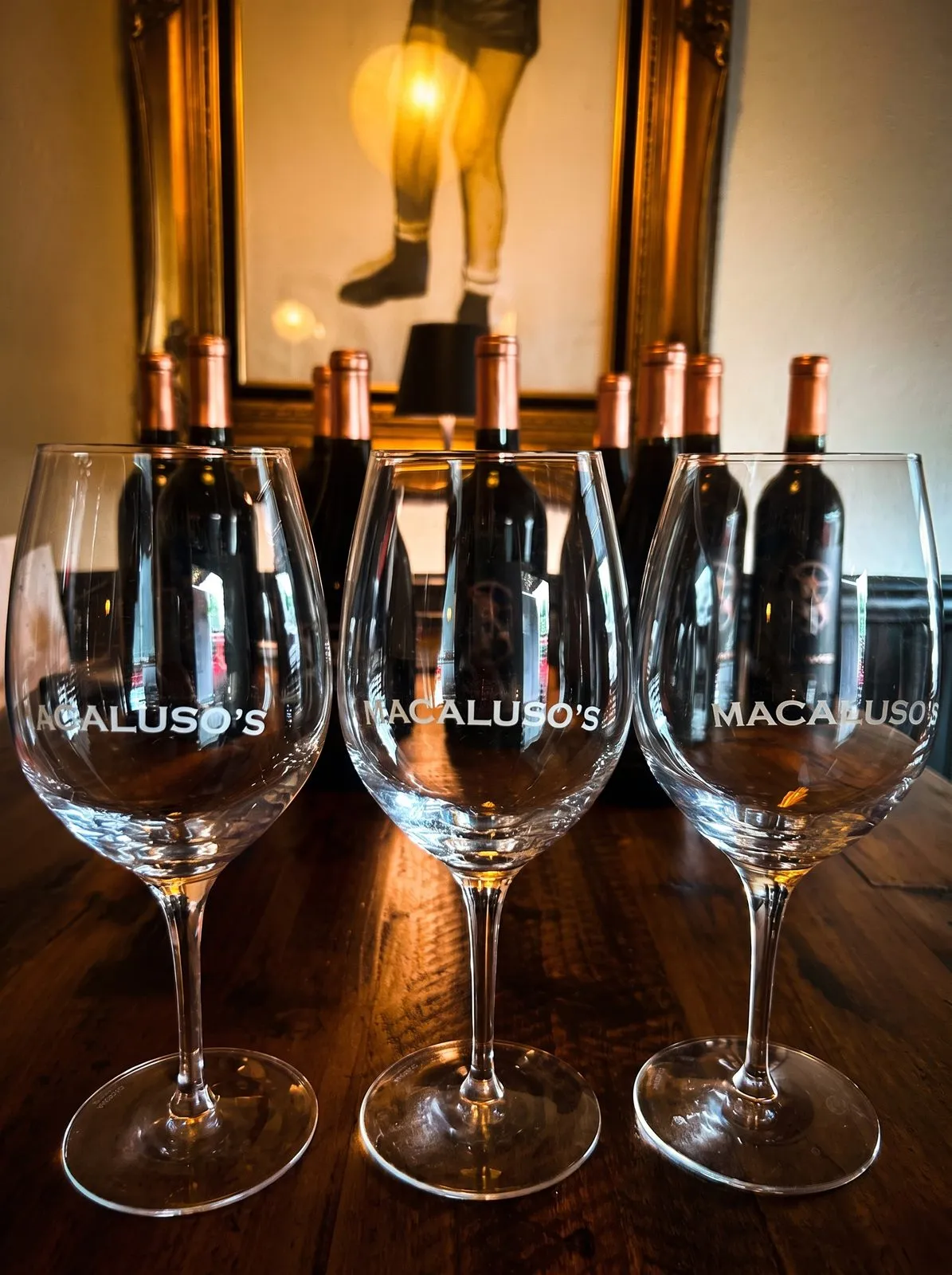 Three Macaluso’s wine glasses on the bar, bottles behind them in warm light.